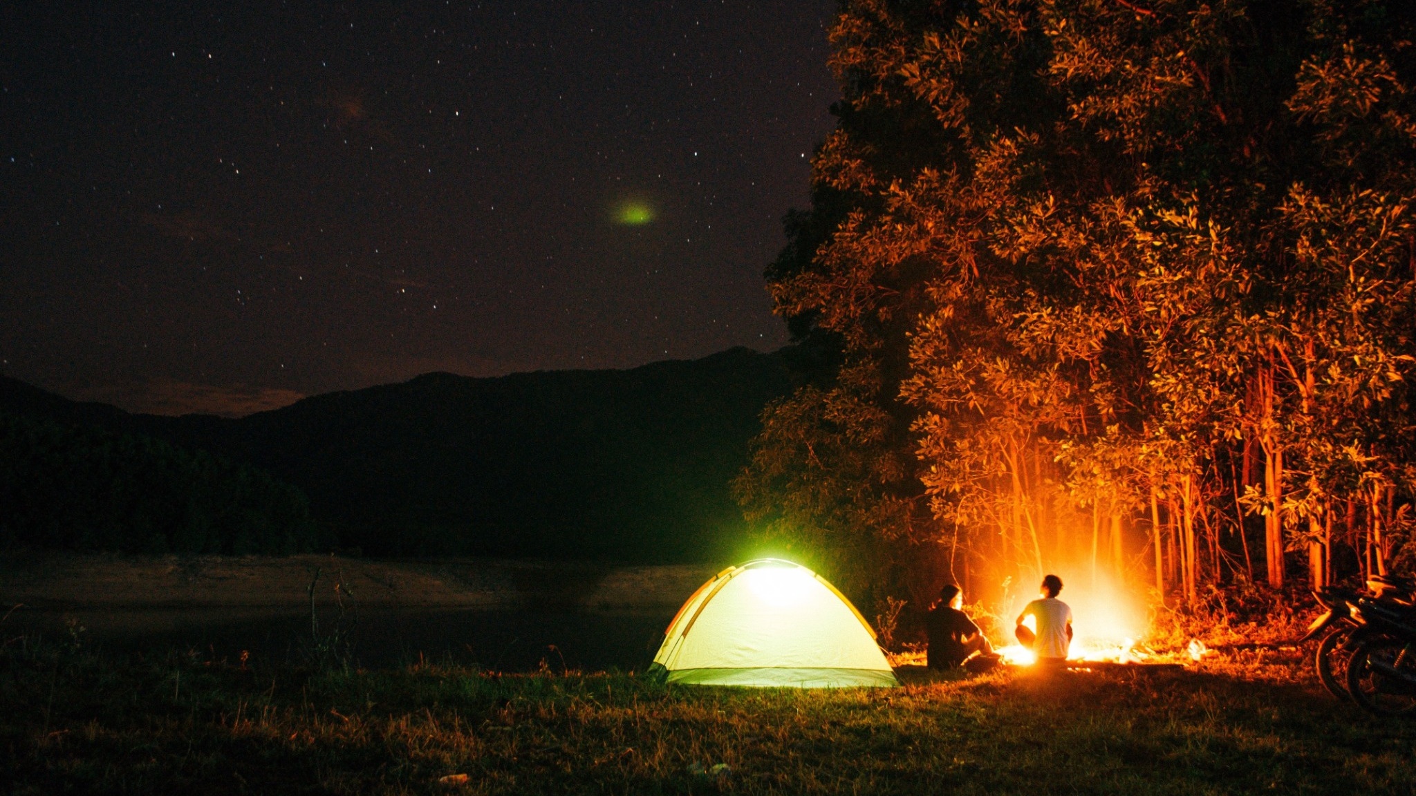 A lit-up dome tent pitched on a lakeshore on a starry night, with a campfire burning beside it; one of the set-ups where the risk of condensation in the tent is highest