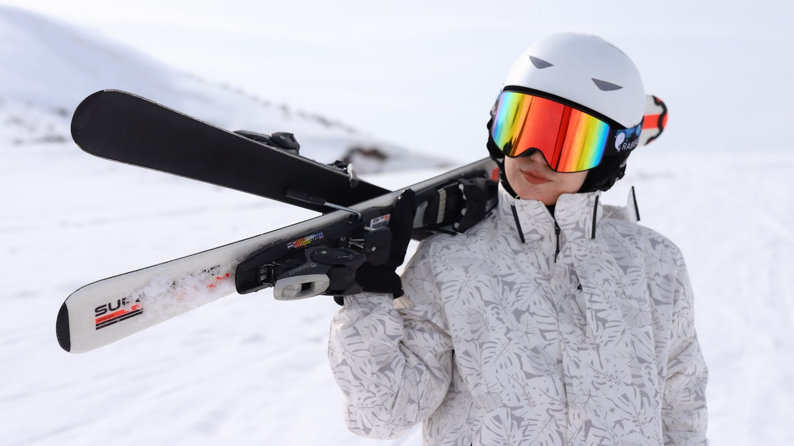 Skifahrer mit weißem Helm, verspiegelter Skibrille und gemusterter Skijacke trägt seine Ski auf der Schulter