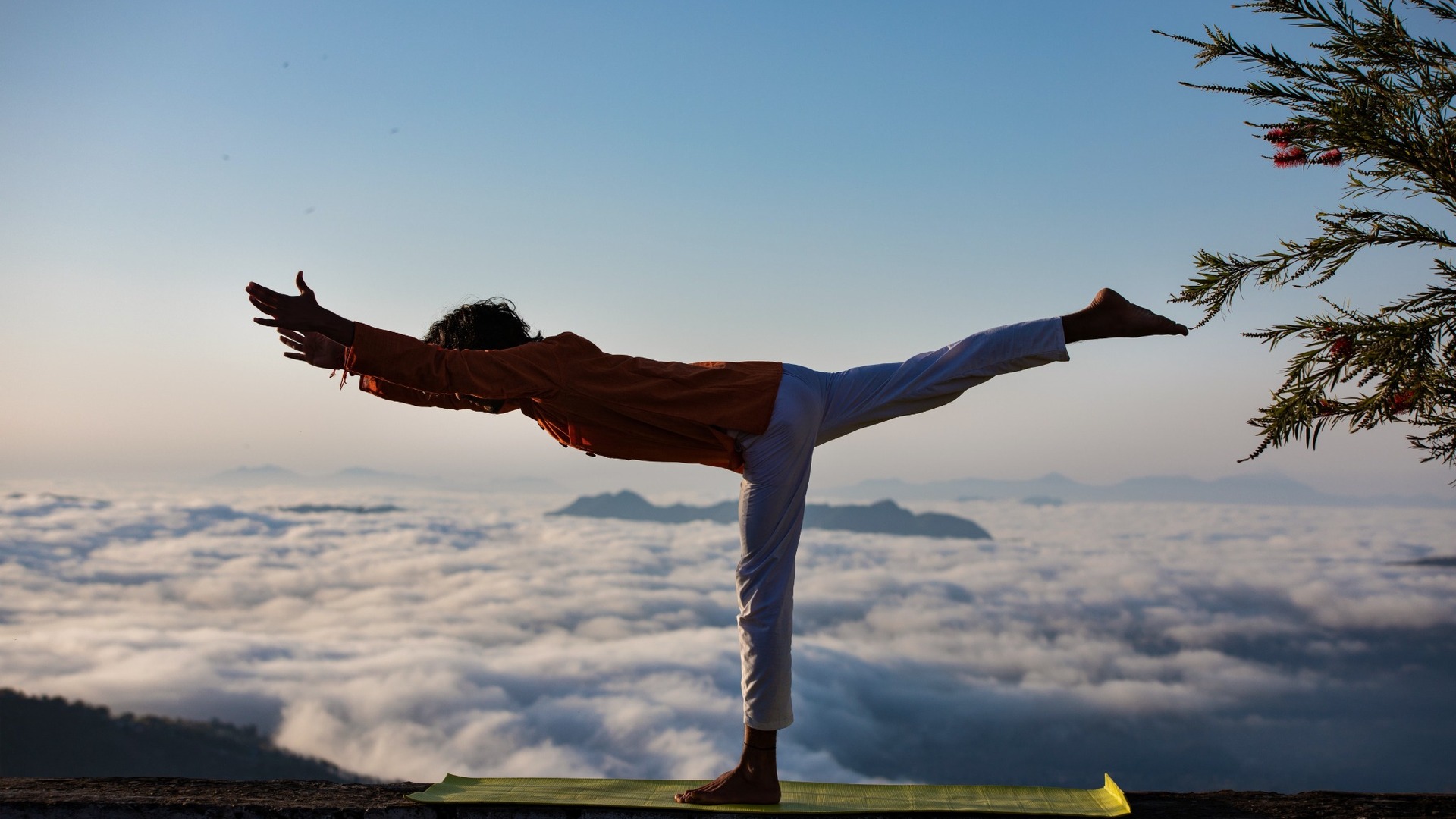 Bulutların üzerinde bir tepede savaşçı pozunda yoga yapan kişi; yoga mı Pilates mi sorusunda yoga tarafını denge, esneklik ve nefes belirliyor