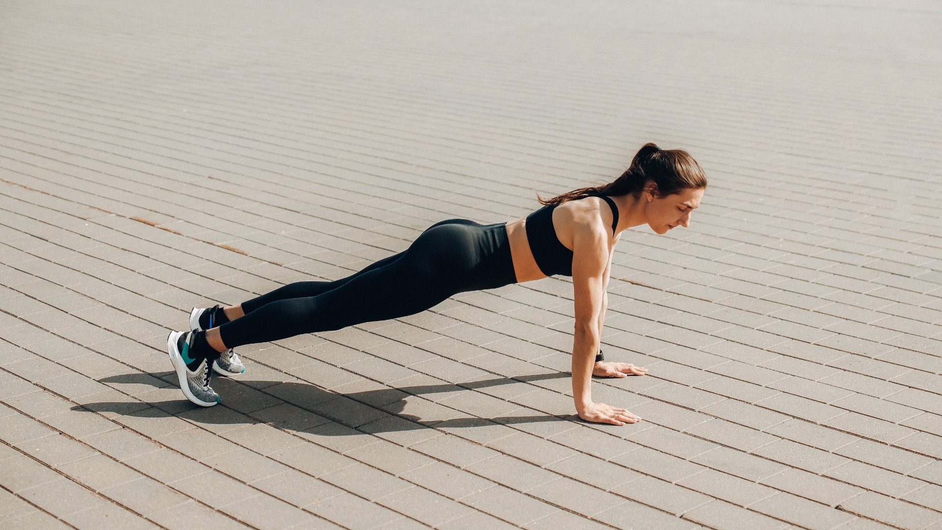 How to do a burpee: a woman holding a straight-arm plank on paving, keeping a straight line from head to heels
