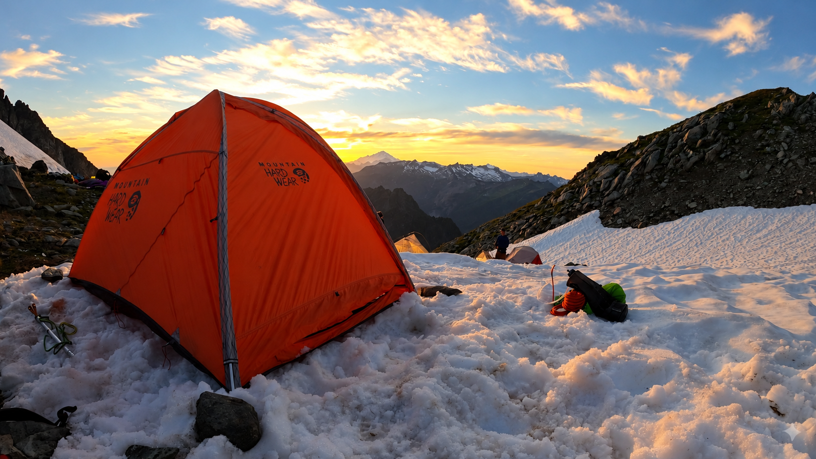 Orange four-season tent pitched on a snowy mountain slope at sunset, with snow-capped peaks behind