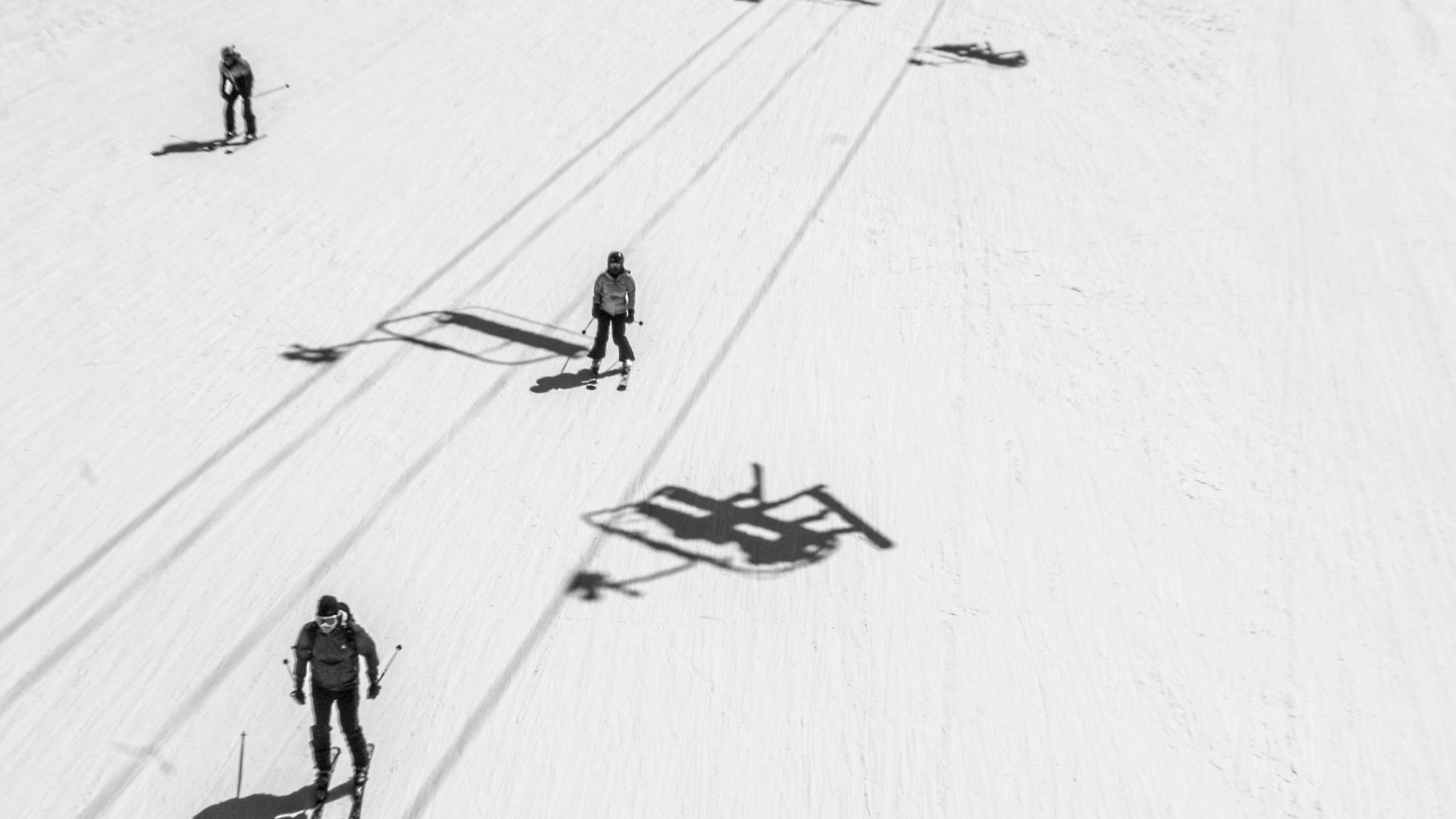 Skiers heading down a wide ski slope crossed by chairlift shadows, black and white