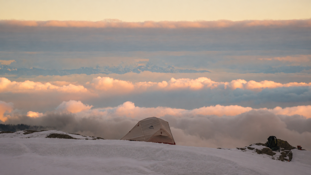 Light-coloured dome tent pitched on a snowy summit above the clouds at sunset