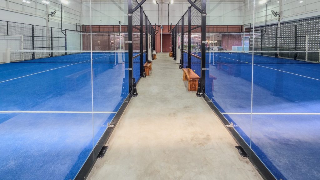 Kapalı tesiste yan yana sıralanmış cam duvarlı padel kortları; padel Americano etkinliğinde her korta dört oyuncu çıkar