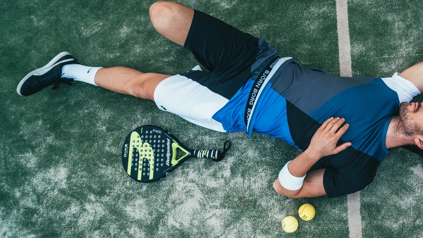 Padel kortunda yere uzanmış oyuncu, yanında padel raketi ve iki top; padel Americano turları arasında kısa bir mola