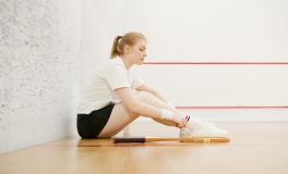 Beyaz duvarlı squash kortunda raketini yanına bırakıp ayakkabısını bağlayan kadın oyuncu