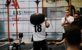 HYROX tarzı bir yarış alanında omzundaki kum torbasıyla sandbag lunge istasyonuna başlayan 18 numaralı sporcu