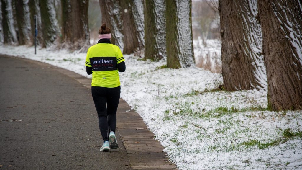 Laufen bei Kälte: Frau läuft auf einem verschneiten Weg zwischen Bäumen mit langärmeliger Basisschicht, Stirnband und langen Lauftights