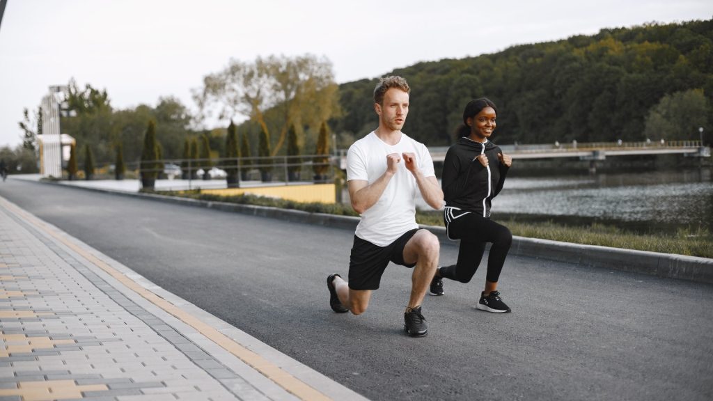 First step in sandbag lunge preparation: a man and a woman doing bodyweight walking lunges on a park path