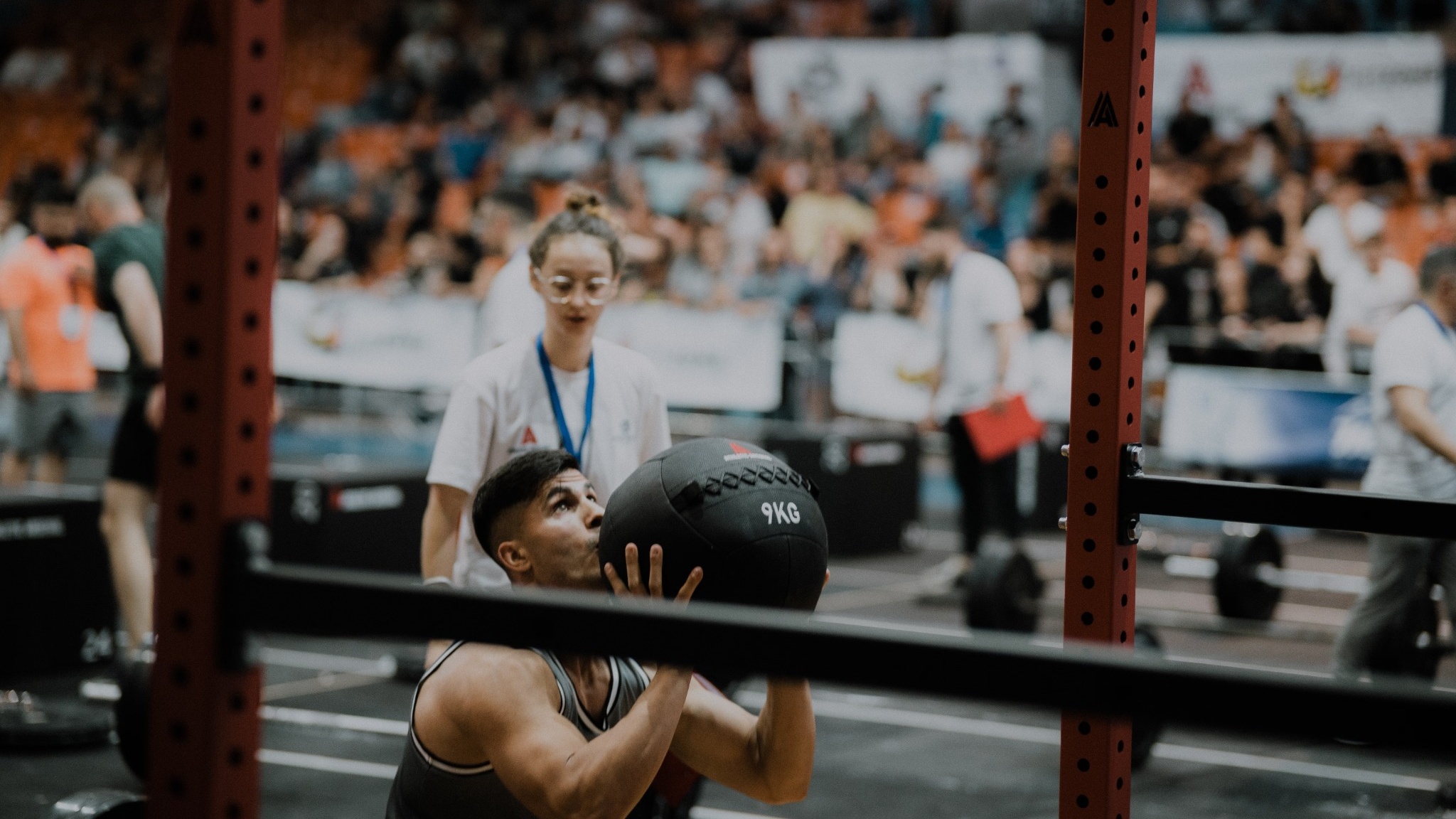 Athlete doing wall balls with a 9 kg ball under a judge’s eye at a competition