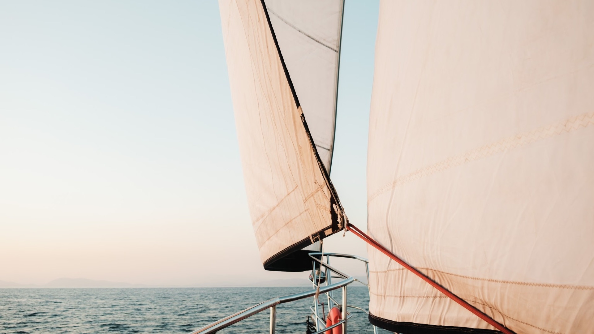 Two white sails filled with wind, seen from a sailing boat’s bow at sunset over a calm sea